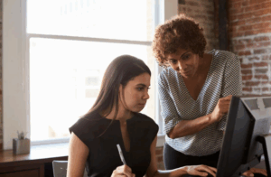Deux femmes collaborant devant un ordinateur, illustrant le mentorat au féminin dans un environnement professionnel masculin, où l’une guide l’autre avec soutien.